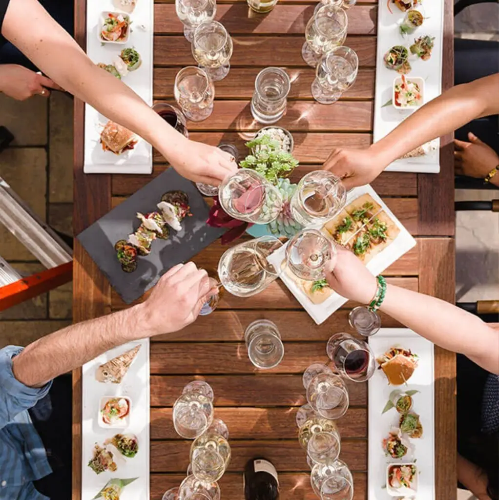 Overhead shot of people clinking glasses over a table filled with appetizers and shared plates at a lively gathering.